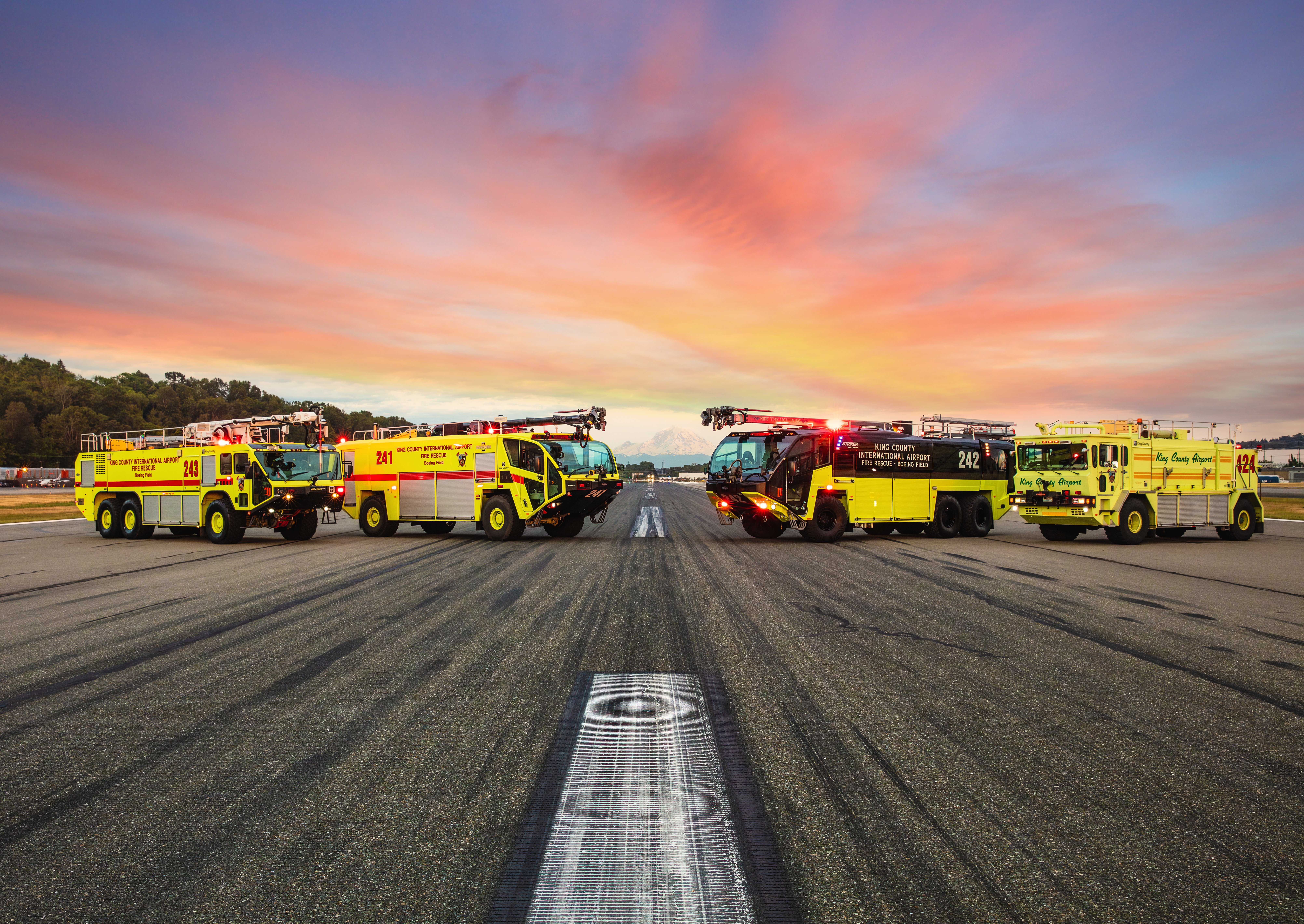 Four Oshkosh ARFF Vehicles parked outside on the runway at sunset.