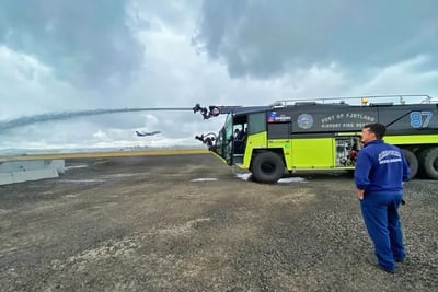 An ARFF firefighter is standing near a Striker 6x6 ARFF spraying water outside on a cloudy day.