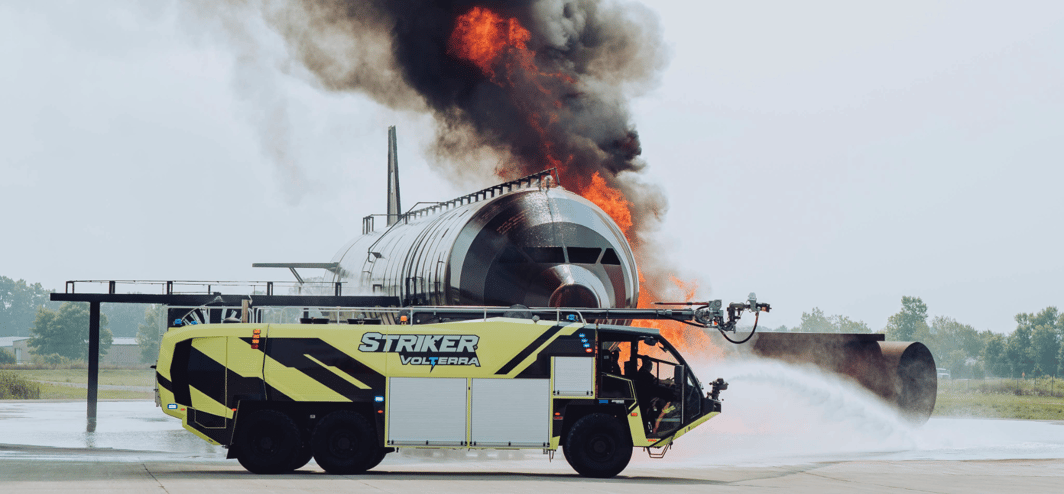 Oshkosh Striker Volterra ARFF vehicle discharging firefighting agents onto a burning aircraft fuselage.