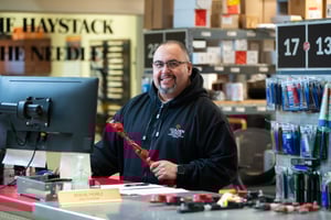 A man in a black Golden State Fire hooded sweatshirt holds a fire truck part behind a counter with a computer screen on it.