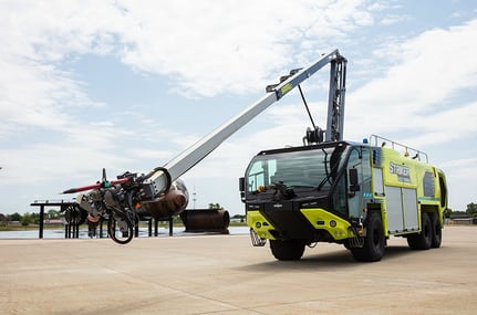 Striker Volterra electric ARFF with the Snozzle high reach extendable turret extended outside on a cloudy day near a demo plane.