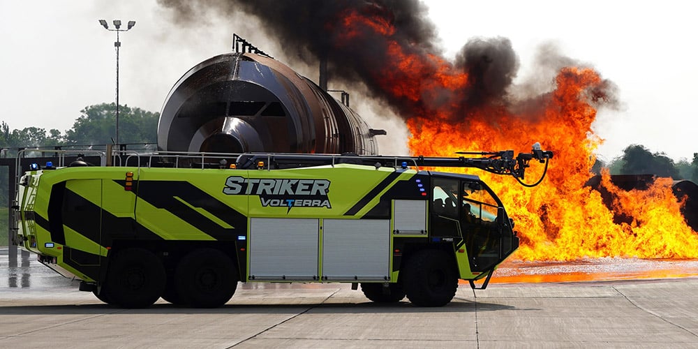 Right side view of Striker Volterra electric Oshkosh Striker ARFF truck driving near a large fire