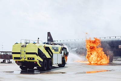 Yellow Striker ARFF truck spraying water on fire that has engulfed a test airplane.
