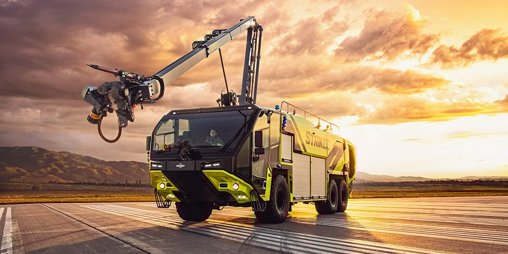 Striker 6x6 ARFF truck with the Snozzle high reach extendable turret and piercing tip extended parked on an airport runway on a cloudy day near mountains.