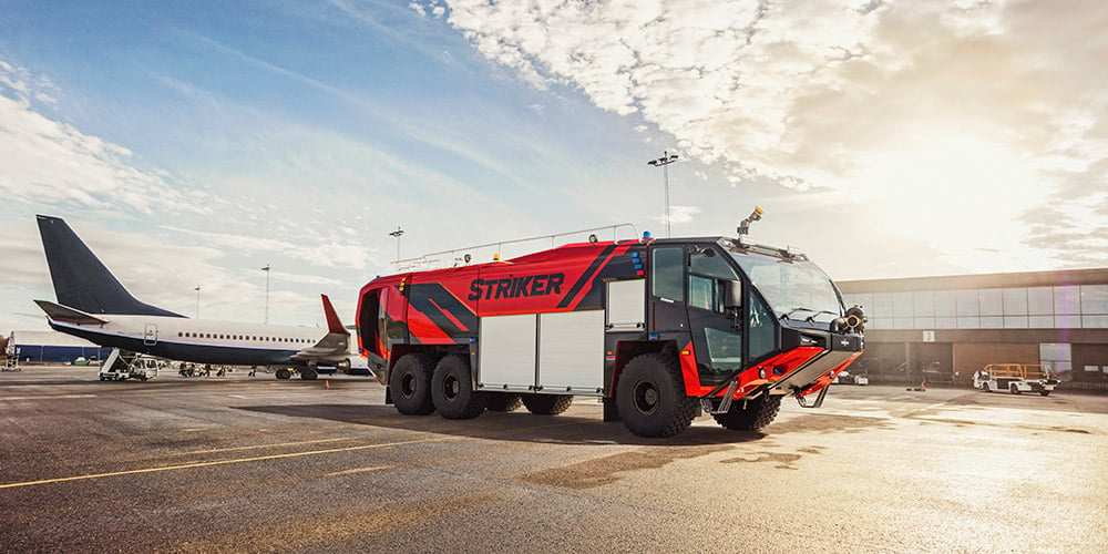 Red Striker ARFF truck parked on airport tarmac with plane and terminal in background.