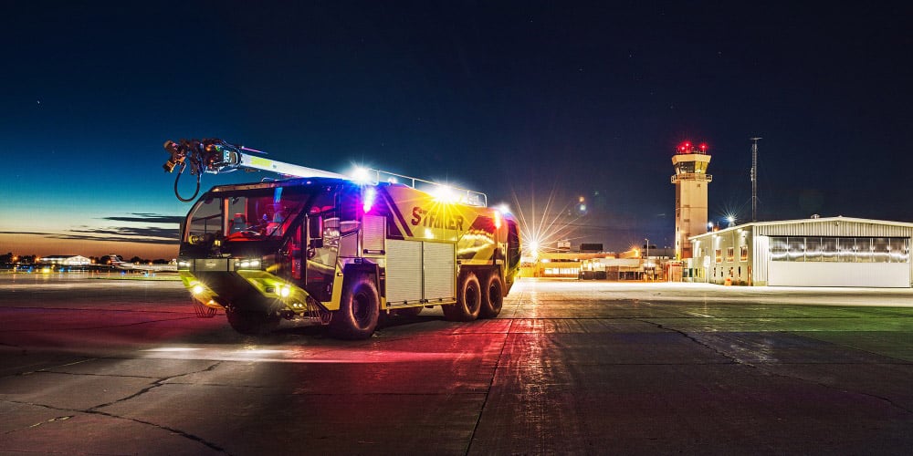 Nighttime shot of yellow Oshkosh Striker ARFF truck with lights flashing sitting on airport tarmac with control tower in the background.