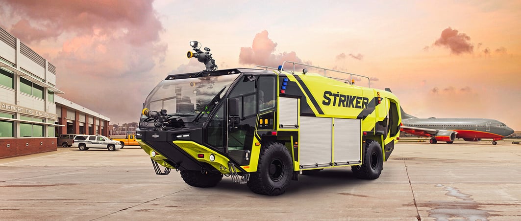 A neon green 4x4 Striker at an airport with airplanes and a building in the background.
