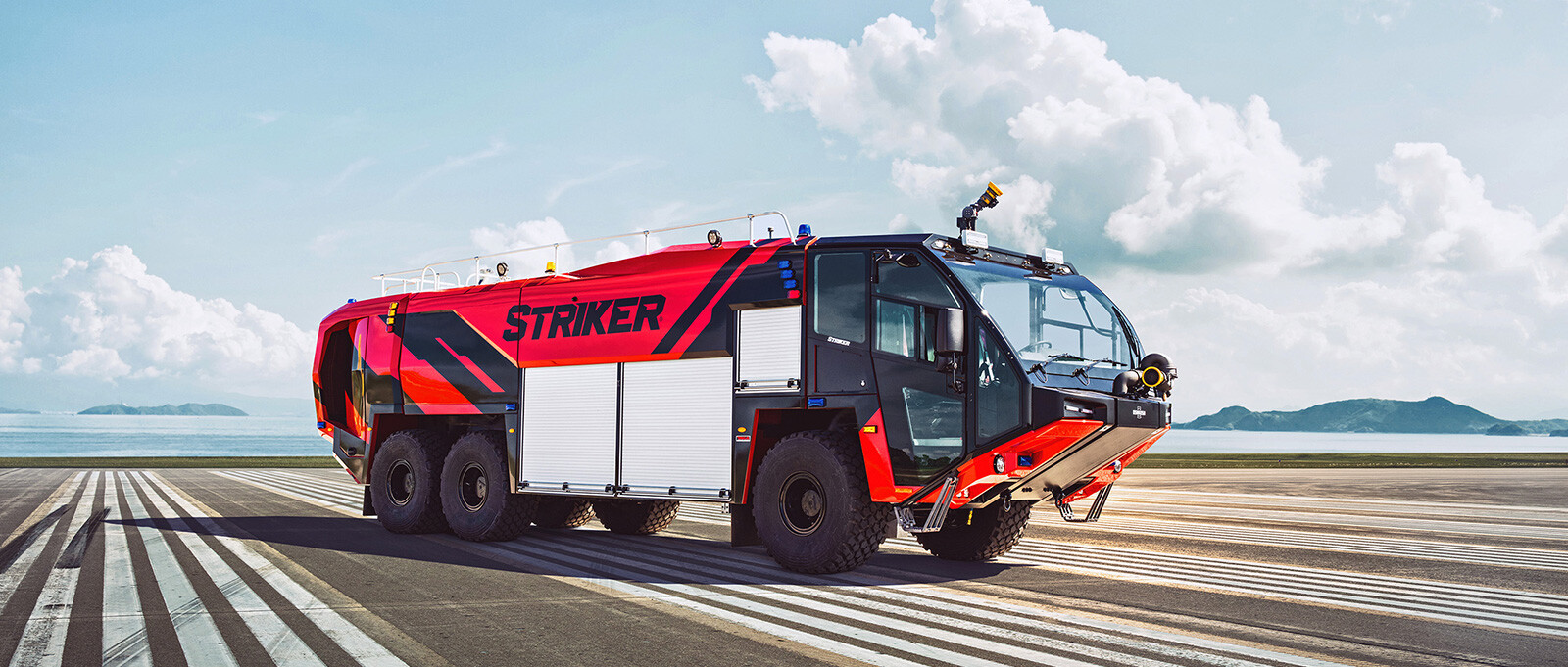 A red 6x6 Striker on a lined pavement patch with a lake and blue sky in the background.
