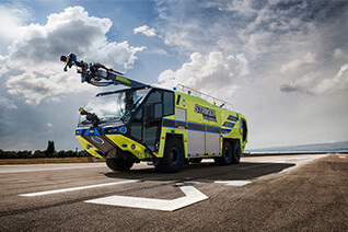 Striker Volterra electric ARFF with a Snozzle high reach extendable turret parked outside on an airport runway on a mostly cloudy day.