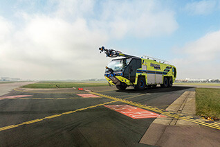 Striker Volterra electric ARFF with a Snozzle high reach extendable turret parked outside on an airport runway on a cloudy day. 
