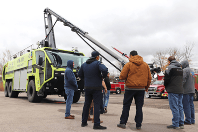 A group of men in winter coats stand outside of an Oshkosh ARFF vehicle with a gray cloudy sky in the background.