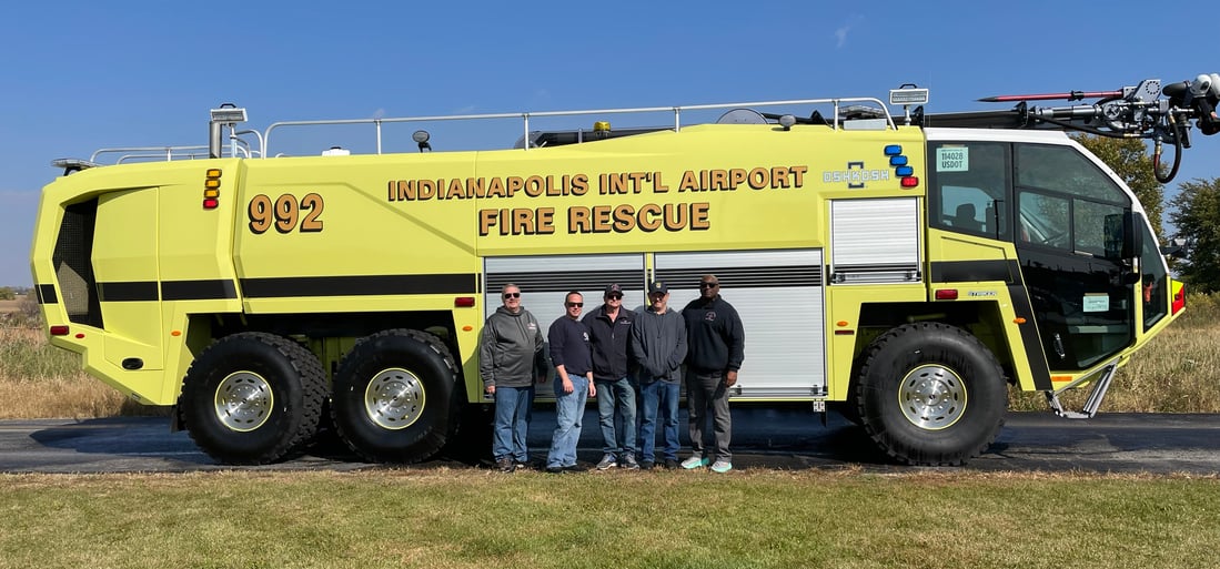 A yellow fire truck sitting on a road near grass, with 5 men in front of it