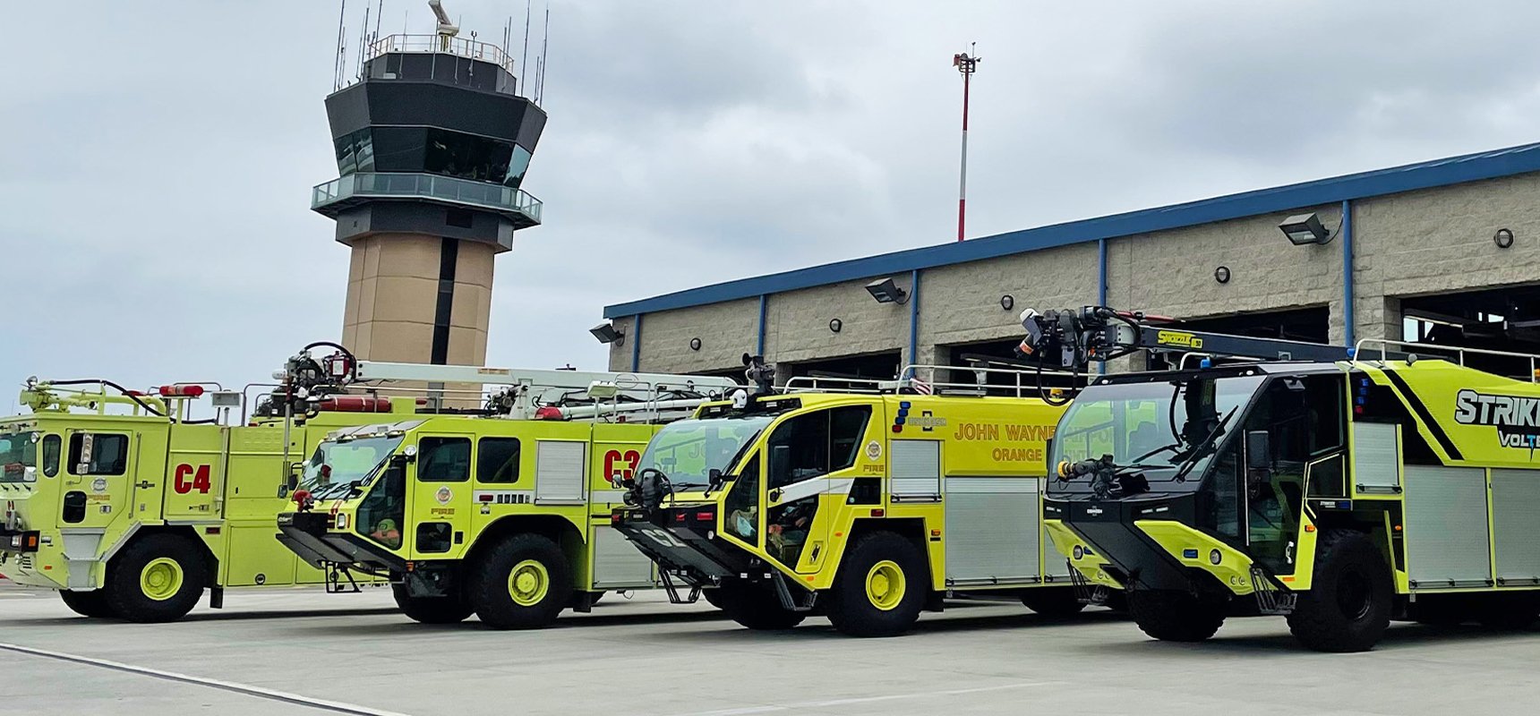 Four neon green trucks, designed for the aircraft rescue and firefighting (ARFF) industry, lined up in front of an airport control tower