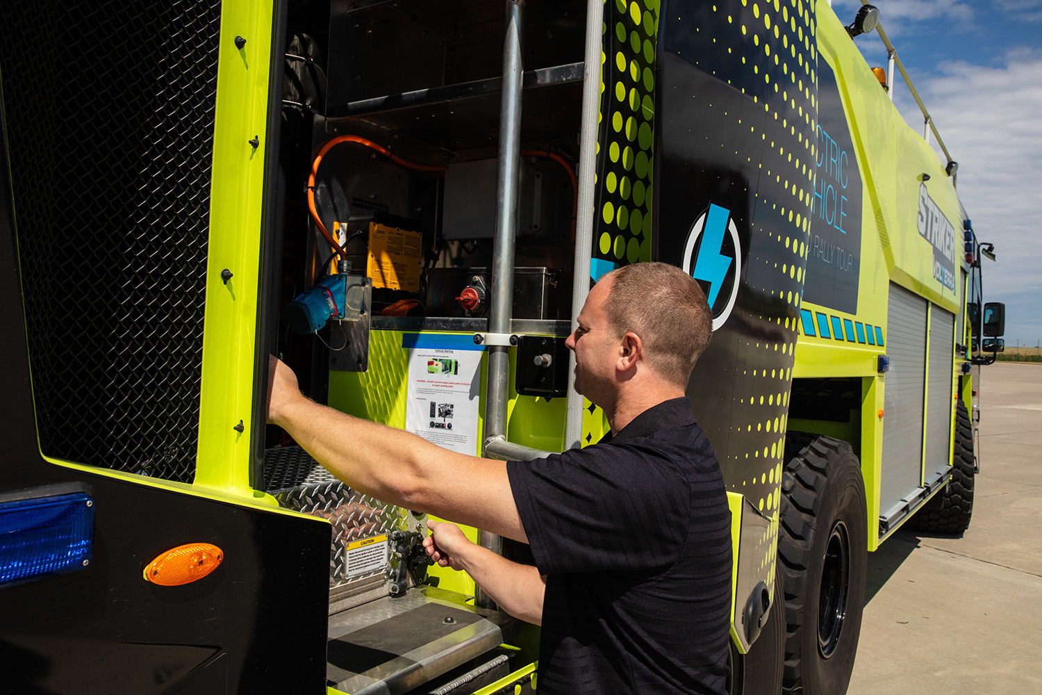 A man wearing a black shirt servicing the engine on a Striker Volterra electric ARFF vehicle parked outside on a sunny day.