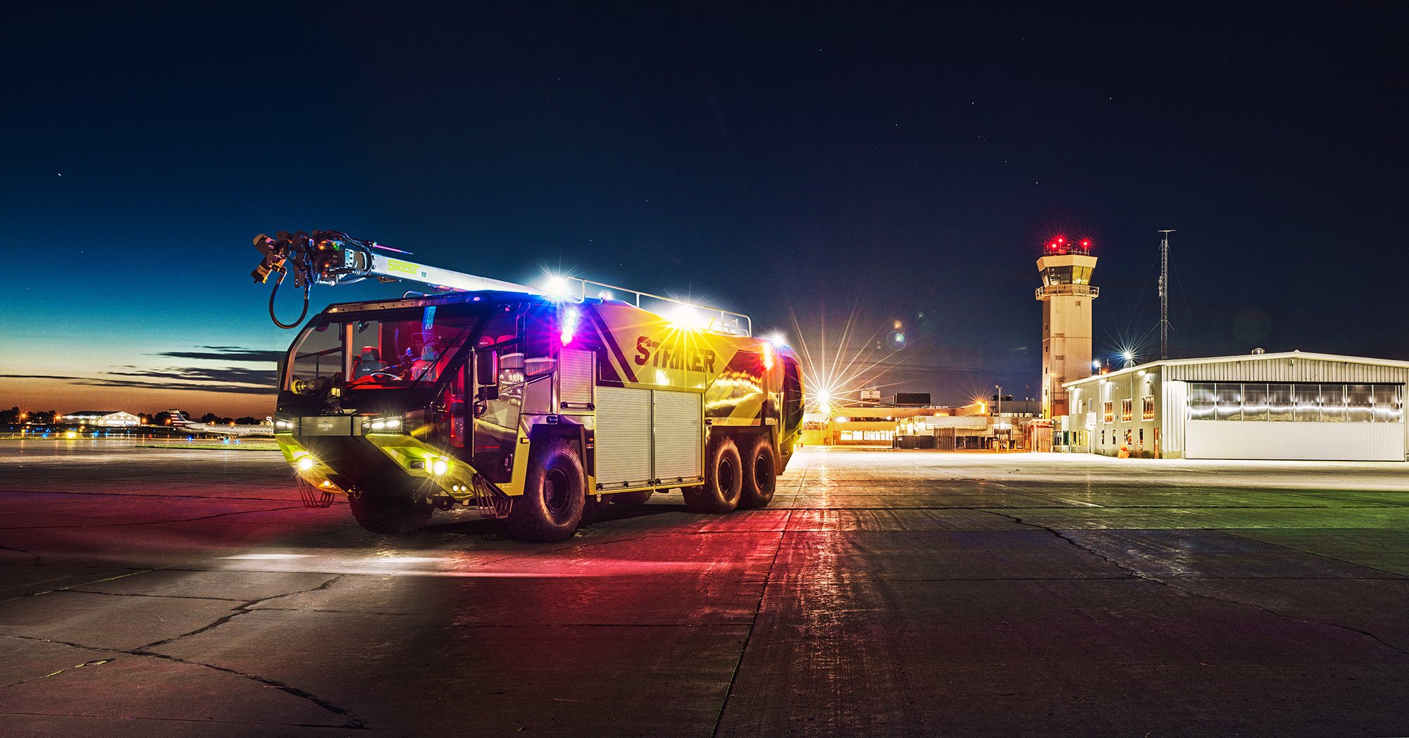Oshkosh Airport Products Striker ARFF with a Snozzle parked outside near an airport at night with its lights on.