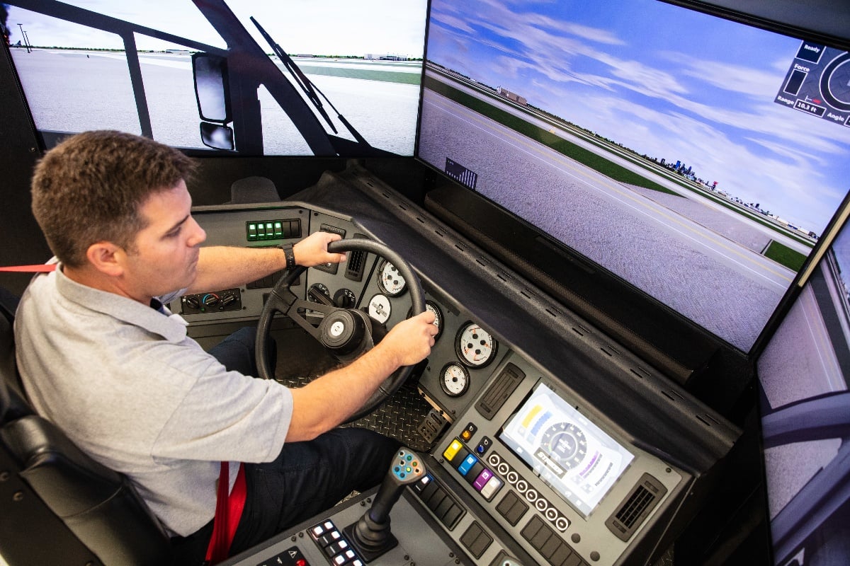 A man sitting in a Striker ARFF simulator chair holding the steering wheel and looking at television monitors with an airport runway. 