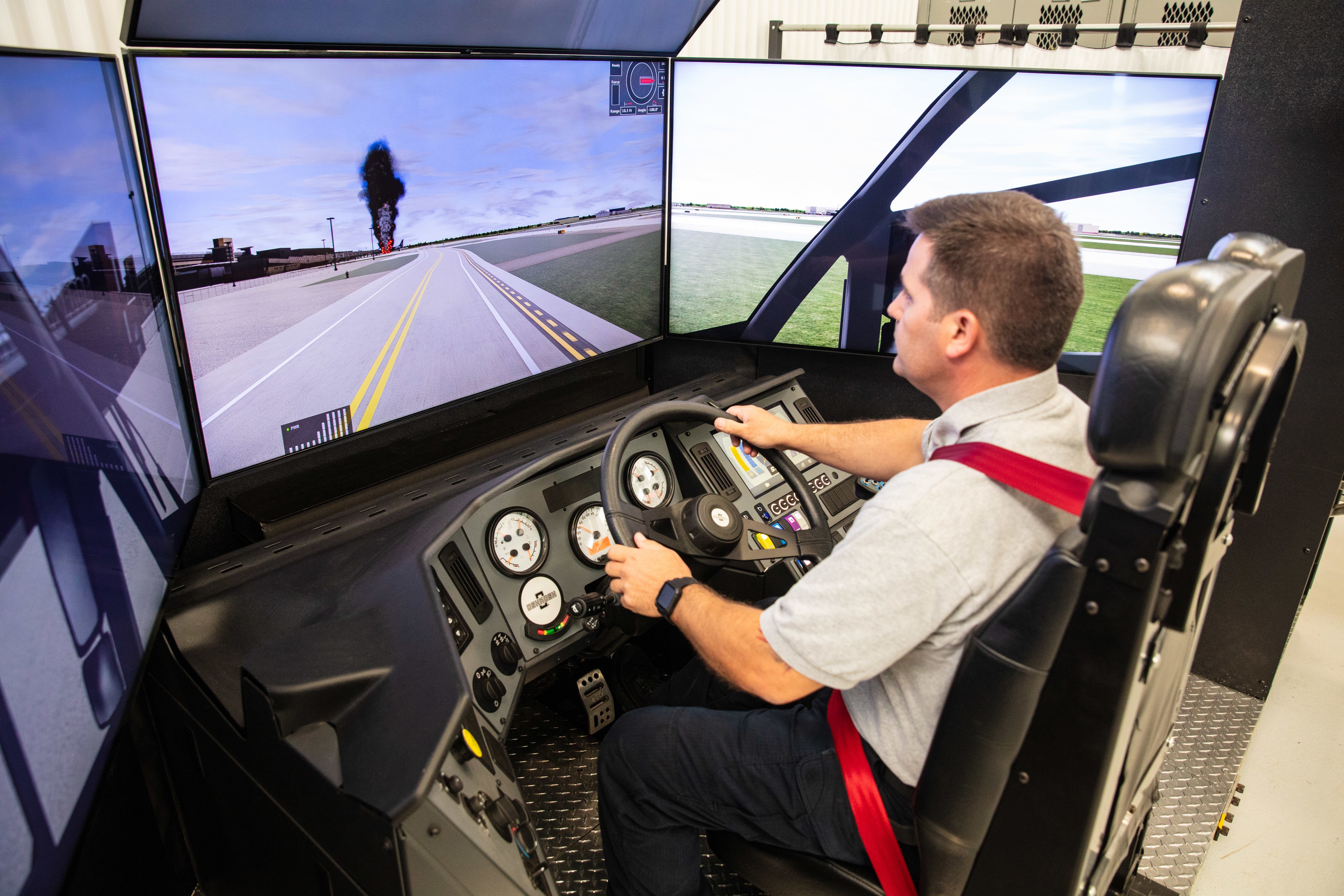 Man sitting in the driver's seat of an Oshkosh Striker ARFF vehicle simulator surrounded by screens.