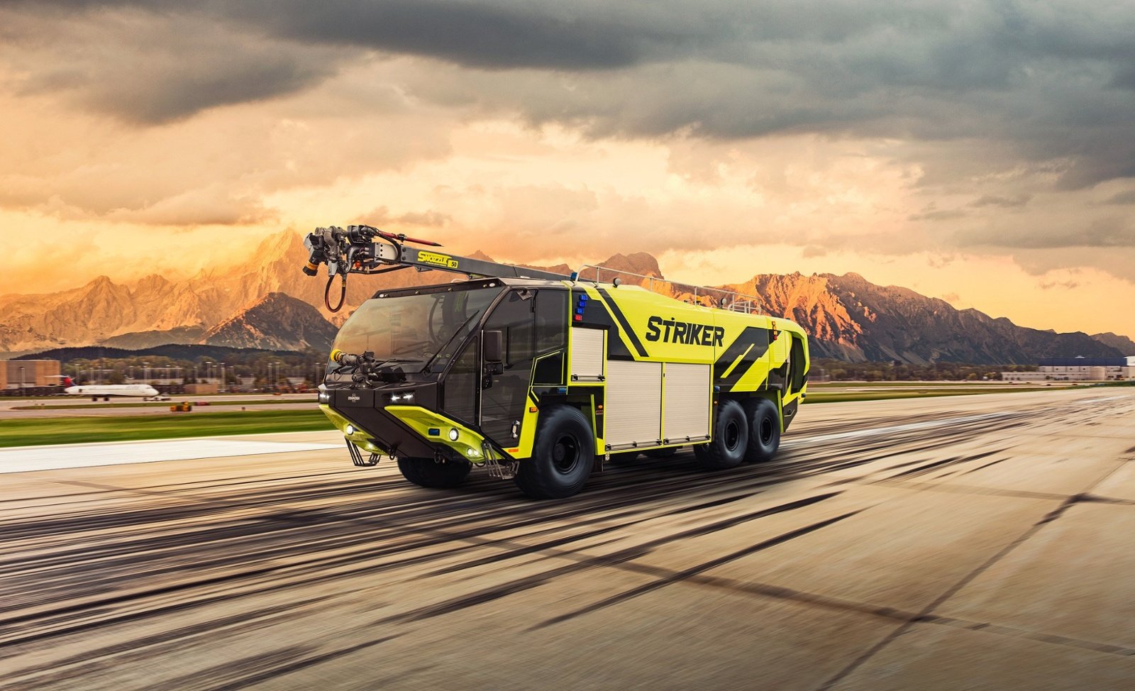 A Striker 6x6 ARFF with a Snozzle high reach extendable turret parked outside on an airport runway near mountains on a cloudy day. 