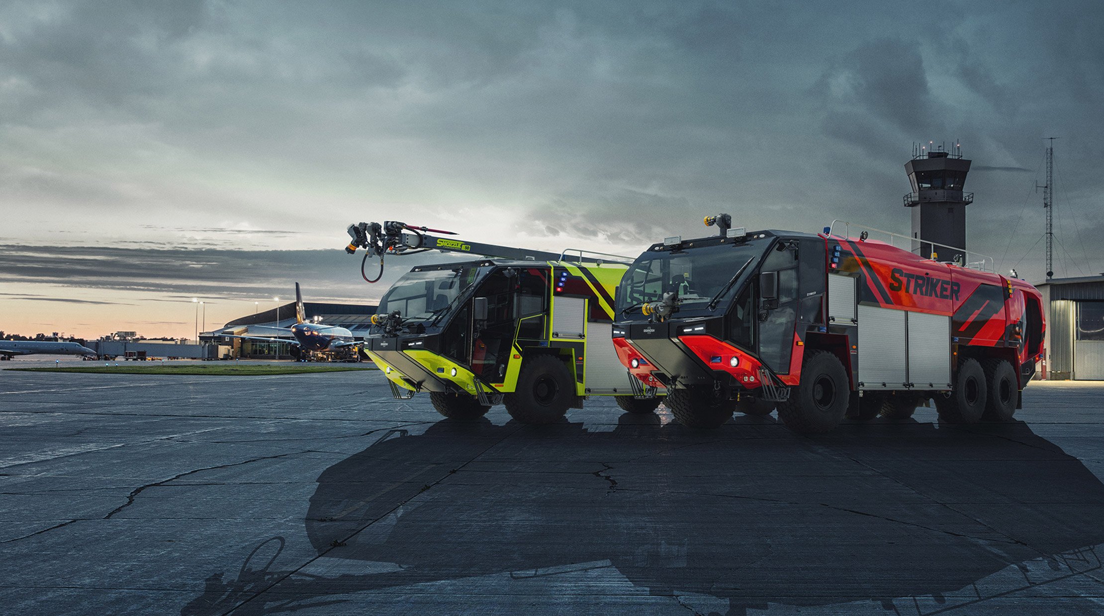 One yellow Oshkosh Striker ARFF and one red Oshkosh Striker ARFF vehicle parked next to each other on the tarmac of an airport in front of a control tower.