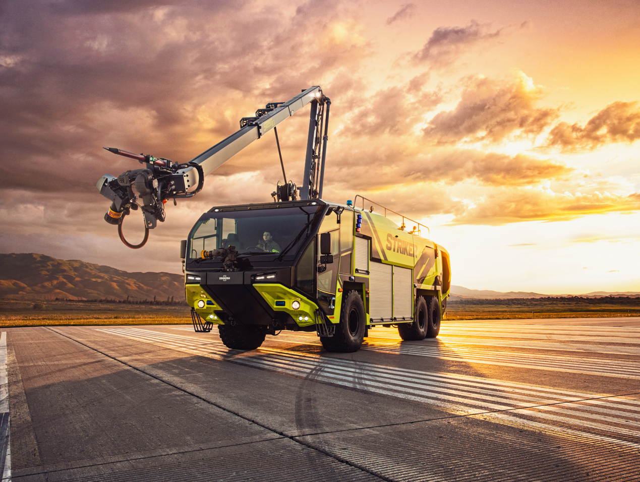 Oshkosh Striker with the Snozzle high reach extendable turret extended forward parked outside on an airport runway on a cloudy day near mountains.